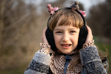 A portrait of a seven-year-old girl in soft warm headphones, in a dress with polka dots and in an autumn coat, listening to music on the street.
