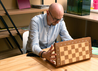 Young bald bearded thoughtful man open box with chess pieces on table. In finding solution, on. Thinking about strategy