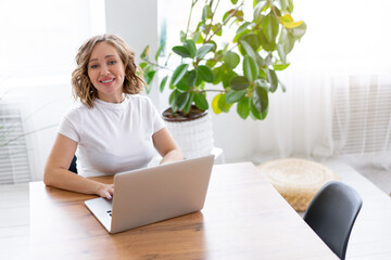 Business woman using laptop sitting near desk white office interior with houseplant looking at camera