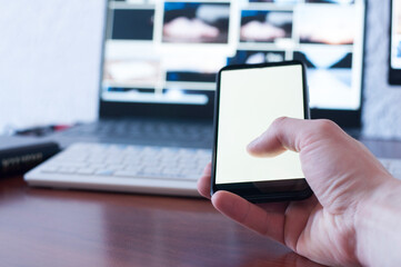 A man businessman is holding a mobile phone, on a wooden table. White blank screen. Empty table. Office workplace. laptop