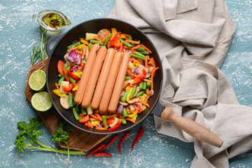 Frying pan with tasty sausages and vegetables on color background