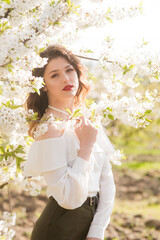 Lovely young girl in a white romantic silk blouse in spring in a blooming cherry garden in sunny weather. Spring and Fashion. Spring blooming gardens