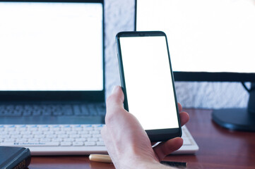 A man businessman is holding a mobile phone, on a wooden table. White blank screen. Empty table. Office workplace.  laptop