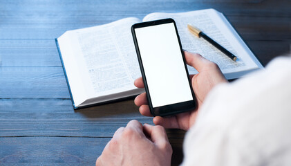 A man businessman is holding a mobile phone, on a wooden table. White blank screen. Empty table. Office workplace. Open book Bible.