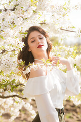 Lovely young girl in a white romantic silk blouse in spring in a blooming cherry garden in sunny weather. Spring and Fashion. Spring blooming gardens