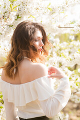 Lovely young girl in a white romantic silk blouse in spring in a blooming cherry garden in sunny weather. Spring and Fashion. Spring blooming gardens