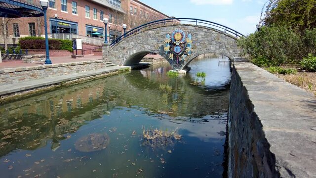 Frederick, MD, USA 04-07-2021: A sunny spring day in the beautiful Carroll Creek Park of Frederick. Static footage shows the stream and sidewalks on both sides with people walking as well as a bridge