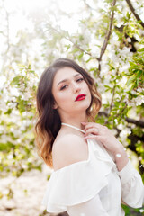 Lovely young girl in a white romantic silk blouse in spring in a blooming cherry garden in sunny weather. Spring and Fashion. Spring blooming gardens