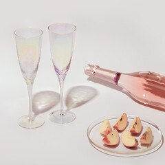 Сlosed bottle of rose champagne sparkling wine and glasses, pieces of apple fruit with sunlight on light table