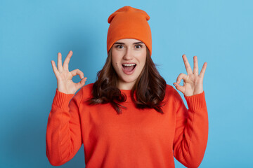 Young European excited female wearing orange sweater and cap looking at camera with opened mouth and showing ok signs with both hands, dark haired girl isolated over blue background.