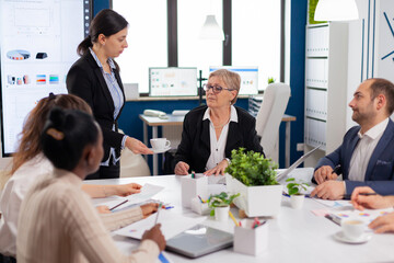 Woman secretary bringing documents and coffee to executive director while multiethnic team planning financial strategy during business conference. Manager briefing teamworkers during brainstorming.