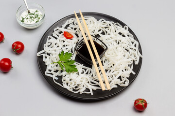 Rice noodles with sauce, bamboo sticks on black plate. Cherry tomatoes in bowl with white sauce on table