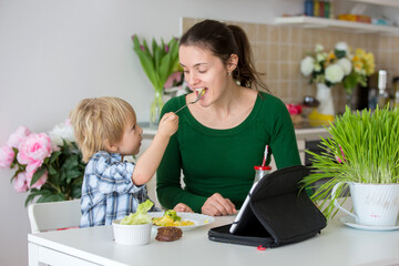 Little toddler child, blond boy, eating boiled vegetables, broccoli, potatoes and carrots with fried chicken meat at home, while watching  movie on tablet, kid giving a bite to mom