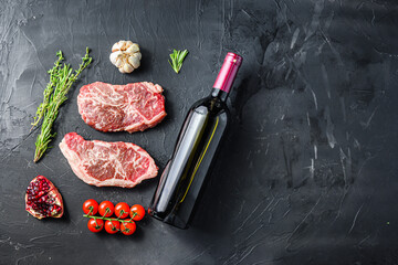 Organic top blade steak cuts, with red wine bottle and glasses, herbs and seasonings on black textured background top view with space for text.