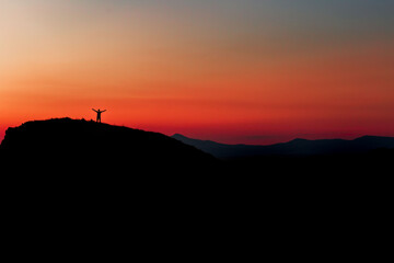 Red sunset with mountain silhouette. A man on mountain in sunset. Traveler on rock waving hands.