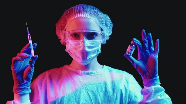 Covid-19 vaccination. Global immunization. Inoculation injection. Female doctor in face mask goggles ppe blue gloves with medication dose vial syringe in red neon light isolated on black background.