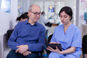 Nurse taking notes on tablet pc discussing with senior patient in dental office, explaining diagnosis. Assistant filling writing healthcare form. Dentistiry hospital.