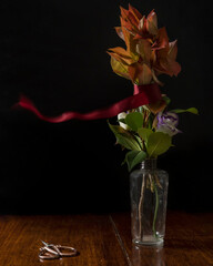 A red ribbon is moved by the wind around a glass vase containing flowers and leaves on a wooden table with vintage copper colored scissors