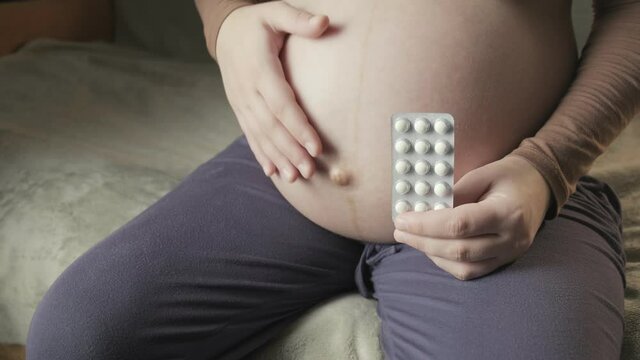 pregnant woman sitting on couch, holding package of pills in palm, strokes belly