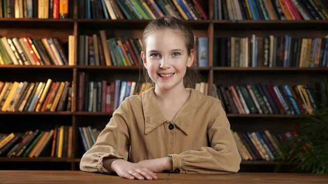 Nice schoolgirl model poses smiling cheerfully while sitting at new brown wooden desk against colourful books on racks and green pot plant