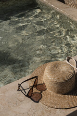Sunglasses and straw hat on marble swimming pool side with clear blue water with waves sunlight...