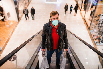 A modern man with bags in his hand and a medical face mask on the escalator in a shopping mall. Shopping during the COVID pandemic - 19 coronavirus