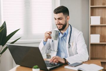 Young male doctor drinking coffee during break and communicating using laptop in his office