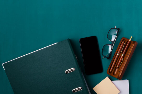 Top view of folder, smartphone with blank screen, glasses, paper for notes, wooden box with pens on the dark green desk