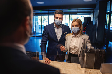 Business couple with face masks standing at reception and checking in the modern hotel. Business trip, relationship, travel during corona, COVID 19 situation, stay safe, keep social distance
