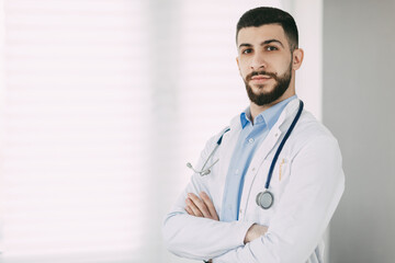 Serious young doctor in stethoscope and white medical coat stands in his office with arms crossed
