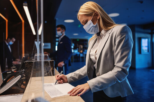 Blond businesswoman with face mask standing at reception in hotel and filling up the form during corona virus pandemic. Business trip, travel during corona, COVID19 precautionary measures