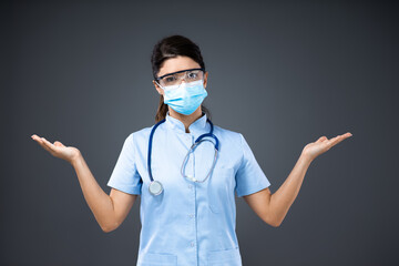 Young confused female doctor with face mask and protective glasses standing with opened hands and looking at camera during corona virus.