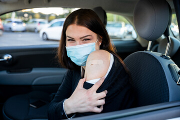 Portrait of a young woman in car shows arm with patch, smiling after having the vaccine against Coronavirus Covid-19 immunizing in the forecourt of the hospital - Person bares shoulder after injection - obrazy, fototapety, plakaty