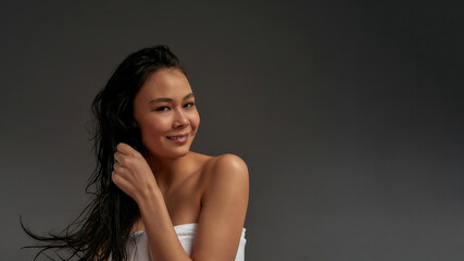 Young beautiful asian half naked woman tousled her hair with one hand in bath towel looks into the camera