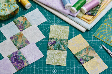 Sewn square pieces of fabric, a stack of fabrics, sewing and quilting accessories lying on a cutting mat
