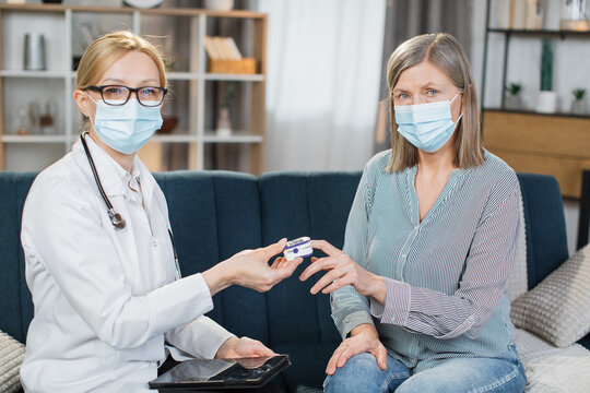 Home Treatment Of Virus. Coronavirus Pandemic. Covid-19 Outbreak. Female Blond Doctor In Face Mask, Examining Sick Patient, Senior Woman, Measuring Saturation Level With Pulse Oximeter.