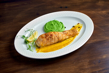 Appetizing grilled salmon steak with yellow sauce and green pea puree, served in a white plate on a wooden background