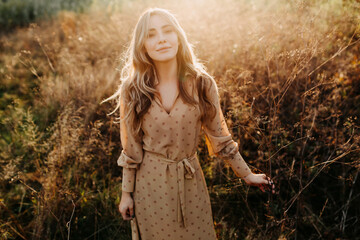 Young woman with long blonde wavy hair, walking in a field with tall dry grass, smiling.