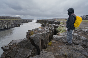 Man in outdoor clothes with yellow backpack standing on a rocky cliff, overlooking the Selfoss waterfall in Iceland.