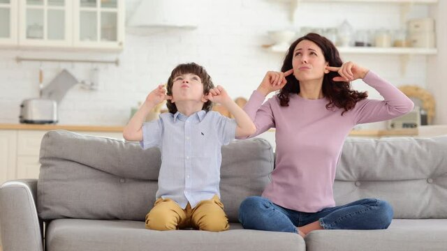 Frowning mom and son sit on couch with closed eyes and cover ears from noisy music or fight sounds from neighbors. Young female parent and kid tired of drilling noise not listen plug ears with fingers