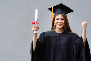 A young happy Asian woman university graduate in graduation gown and mortarboard holds a degree...