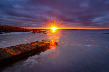 a bright sunset with the sun in a narrow skylight with sunbeams and a dramatic cloudy sky over a frozen lake with an old wooden pier and coastal rope. picturesque winter landscape