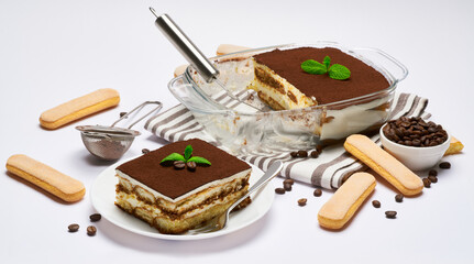 Traditional Italian Tiramisu dessert in glass baking dish, portion on plate and scapula isolated on white background
