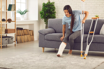 Young smiling woman with broken leg in cast trying to stand up with crutches from sofa at home with...