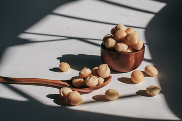 Macadamia nuts in wooden bowl on white table with sunlight and shadow. Macadamia nuts are a source of protein packed with healthy vitamins.