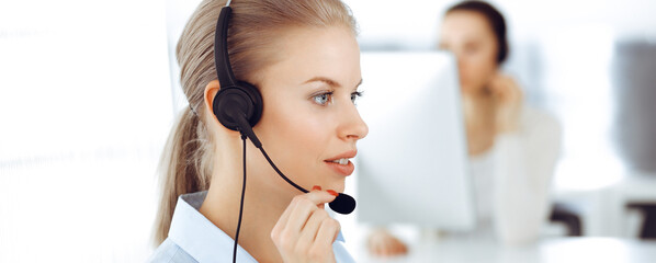 Blond woman call operator is using computer and headset for consulting clients online. Group of...