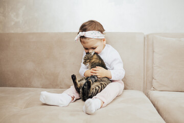 Child girl hug and kiss the his cute funny cat with tenderness and love while sitting on the sofa in the living room.