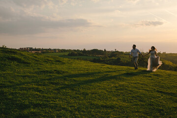 Fototapeta premium Beautiful happy wedding couple of groom and bride holding hands while running on the green grass in the hill in mountains on sunset.