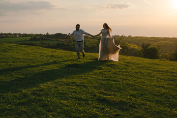 Beautiful happy wedding couple of groom and bride holding hands while running on the green grass in the hill in mountains on sunset.