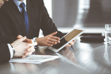 Unknown businessmen using tablet computer and work together at the glass desk in modern office, close-up. Teamwork and partnership concept